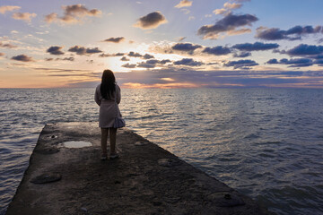 A girl with long black hair in a beige coat stands on the pier and looks at the setting sun. Cloudy sky, calm gray sea.