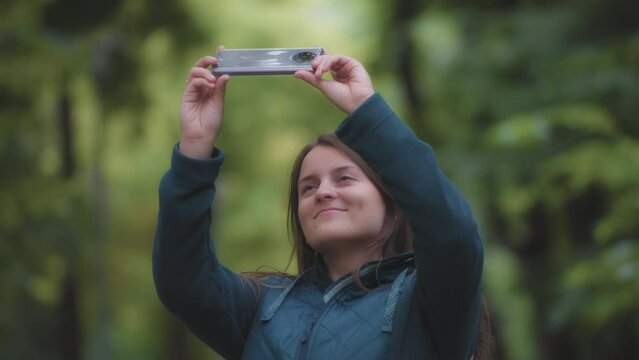 A smiling woman taking pictures of the trees with a phone, blurred green natural background. Slow motion. 