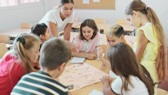 Schoolkids playing interesting board game with teacher in classroom. Boys and girls playing tabletop game in school.
