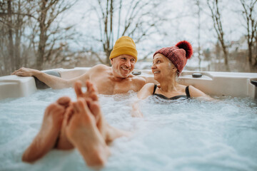 Senior couple in kintted cap enjoying together outdoor bathtub at their terrace during cold winter...