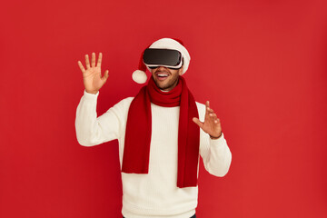 Fototapeta premium Focused Man Trying VR. Portrait of Multiracial Amazed Guy in Santa Hat Discovering New Technologies Wearing Virtual Reality Headset, Futuristic 3d Vision. Indoor Studio Shot Isolated Red Background 