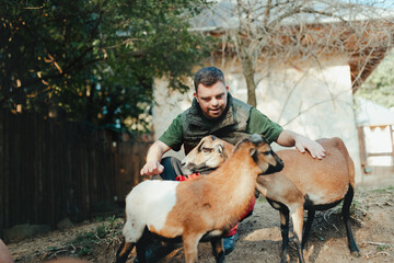 Caretaker with down syndrome taking care of animals in zoo, stroking goats. Concept of integration people with disabilities into society. - obrazy, fototapety, plakaty