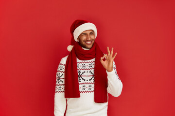 Smiling Man Showing Ok Gesture. Satisfied Multiracial Guy in Santa Hat Making Ok Symbol with Fingers, Approving, Satisfied. Indoor Studio Shot Isolated on Red Background 