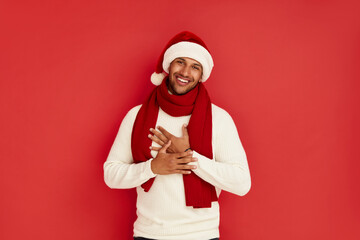 Fototapeta na wymiar Happy Man Smiling Camera. Cheerful Guy in Santa Hat Smiling Sincerely, Holding Hand at Chest, Positive Mood. Indoor Studio Shot, Isolated on Red Background 