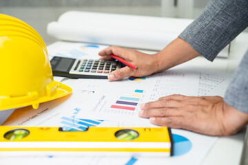 Asian engineer with architectural house plan project blueprint, graph and yellow helmet and engineering construction tools.