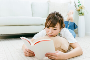 部屋に寝転んで本を読むアジア人女性（読書）
