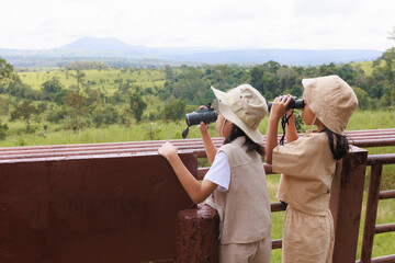 Excited asian child girls looking through binocular searching for animal in savannah green forest....
