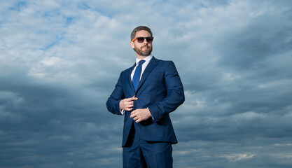 Manager wearing sunglasses sky background. Grizzled manager buttoning suit jacket. Serious manager