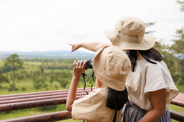 Excited asian kid looking through binocular searching for animal with her mother in savannah green forest. Children learning with nature and travel with family on camping vacation.
