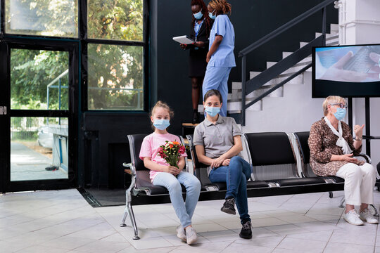 Mother Sitting Beside Child Waiting For Grandfather To Finish Consultation During Checkup Visit In Hospital Area. Girl Holding Bouqet Of Flowers, People Wearing Medical Face Mask Against Covid19