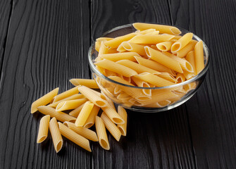 Uncooked penne pasta in glass bowl on black wooden table