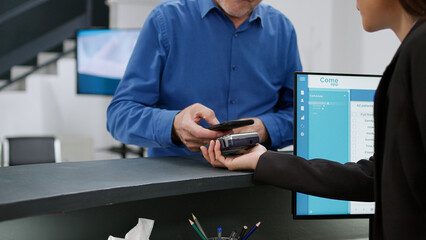 Senior patient paying appointment and medication with credit card at hospital reception counter. Old man making electronic transation and payment for checkup at registration desk. Handheld shot.