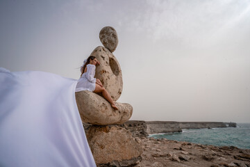 A woman sits on a stone sculpture made of large stones. She is dressed in a white long dress,...