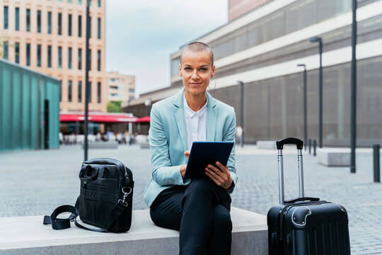 Smiling businesswoman with tablet PC and luggage sitting on bench at footpath