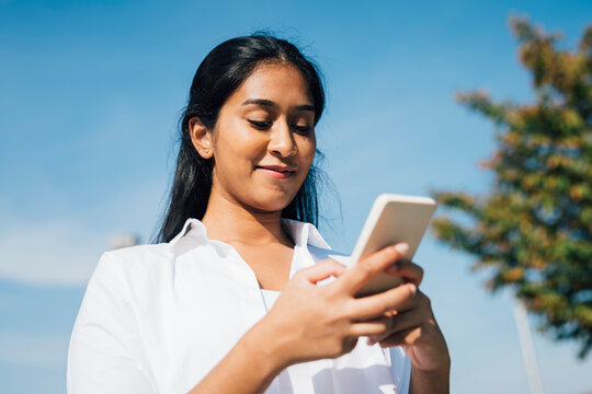 Smiling woman text messaging through mobile phone under blue sky