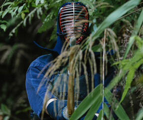 Portrait of kendoka man in the forest holding the sinai-sword behind a plant.  Kendo is the Japanese martial art of sword fighting