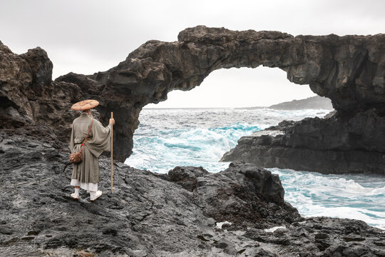 Monk Wearing Hat Standing In Front Of Rock Arch