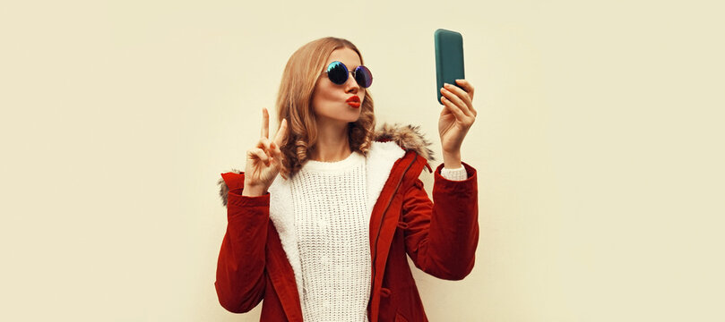 Portrait of beautiful young woman taking selfie with smartphone blowing her lips sends kiss wearing red jacket with fur hood