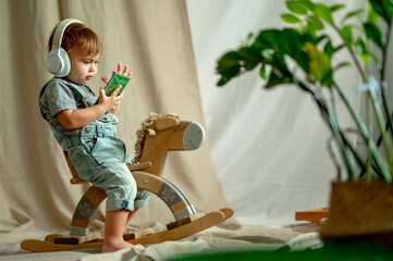 Cute baby boy wearing wireless headphones using smart phone on rocking horse