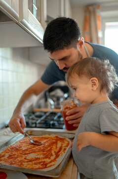 Boy by father spreading tomato sauce on pizza dough in kitchen