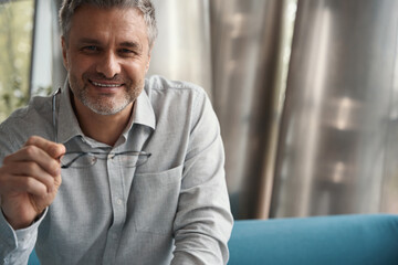 Friendly gray-haired businessman sits on sofa,holds glasses in his hands