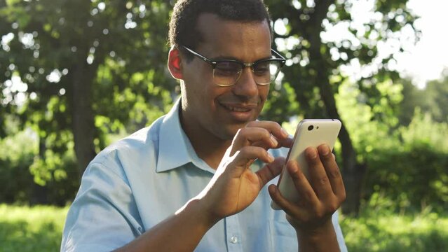 Funny biracial man in eyeglasses dancing and singing aloud listening to music in special music player or radio application on smartphone, scrolling web-pages or watching videos with sound