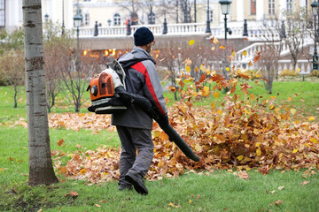 Man worker clean the city street with leaf blower. Falling leaves removal in autumn park