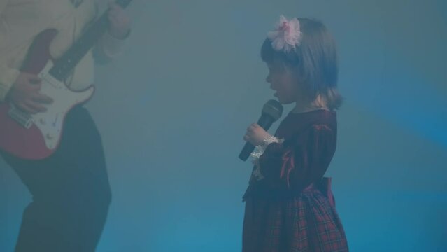 Little girl on stage in vintage dress, she sings into microphone and dances, her father plays an electric guitar. Color music is shining and smoke is billowing. Family time, creativity and hobbies