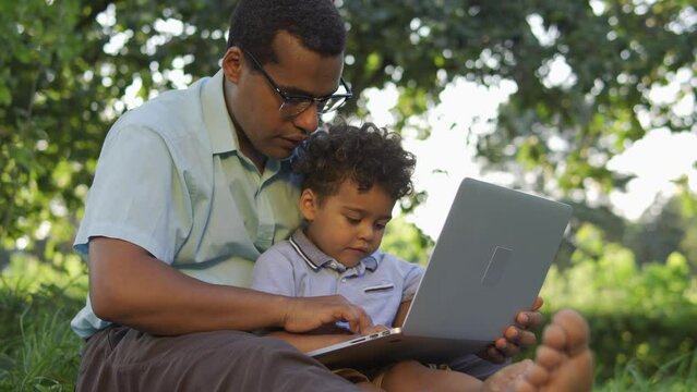 Multiethnic father trying to work on laptop with little curly-haired son sitting nearby and helping, barefoot boys sitting on the grass outdoor, balancing personal life and work, paternity leave