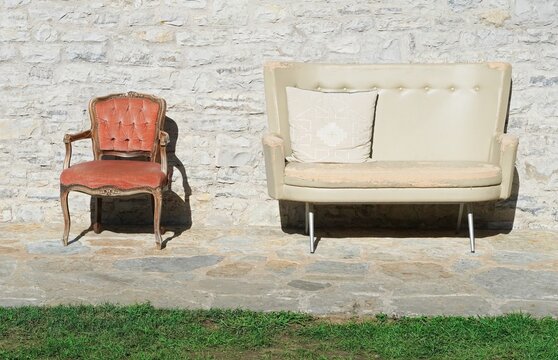 Two chairs in used condition with worn fabric, different styles and padded material.  Empty double seat sofa and armchair near wall with sunlight