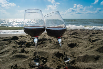 Close-up of two glasses of red wine standing in the sand on the beach against a blue sky with clouds and rays of the sun
