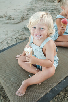 From Above Of Adorable Little Boy With Blond Hair Enjoying Tasty Ice Cream While Sitting On Beach Near Crop Friend