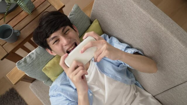 top view of a happy Asian male lying on couch is cheering with raised hand for winning the racing game on the mobile phone in the living room at home