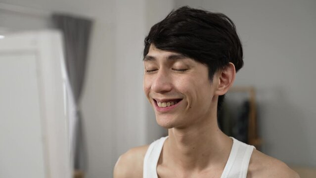 shoulder shot cheerful asian young man in undershirt smiling at his reflection in the mirror feeling positive at the beginning of the day in his bedroom at home
