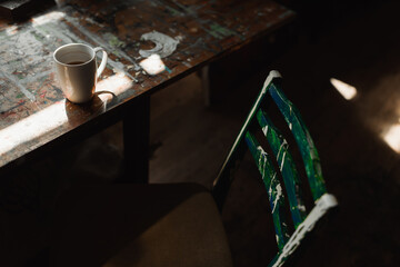 old wooden chair near rough table with paint spots and white cup of tea in art studio.