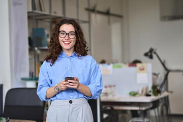 Smiling young business woman, happy businesswoman corporate leader holding cellular smartphone working standing in office using mobile cell phone working on cellphone looking at camera.