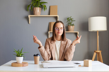Portrait of attractive woman sitting at workplace with laptop and raising hands in mudra gesture,...