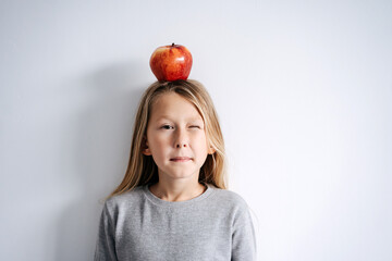 Blinking with one eye boy with an apple on his head. Over white wall.