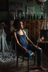 brunette african american woman in overalls sitting in workshop with vintage decor and looking away.