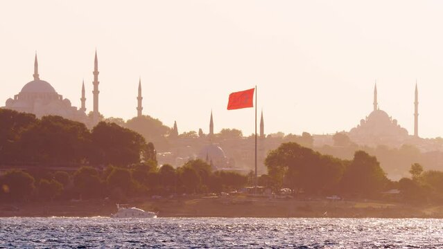 Fatih area of Istanbul seen across Bosporus, telephoto perspective from Asian side. Red flag on tall pole, mosque silhouetted on background. Single yacht slowly cruise on water. Sunny summer evening