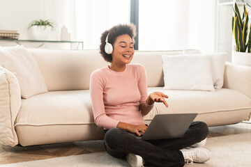 Obraz premium Cheerful millennial african american woman in wireless headphones has video call on laptop, gesturing