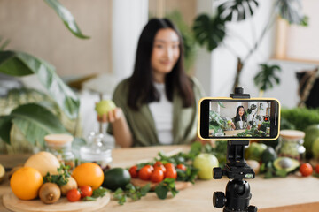 Smiling young asian woman cooking vlogger live-streaming her video blog to followers, while showing...