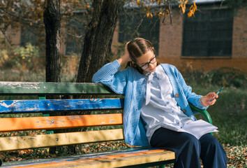 A teenager on a bench is doing homework on a sunny autumn day.