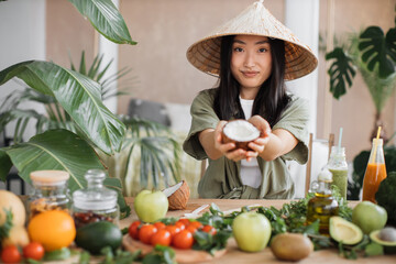 Beautiful asian woman in traditional conical hat holding coconut sitting at table with fruit and vegetables ingredients to make healthy vegan salad, indoor at table at stylish tropical kitchen. - obrazy, fototapety, plakaty