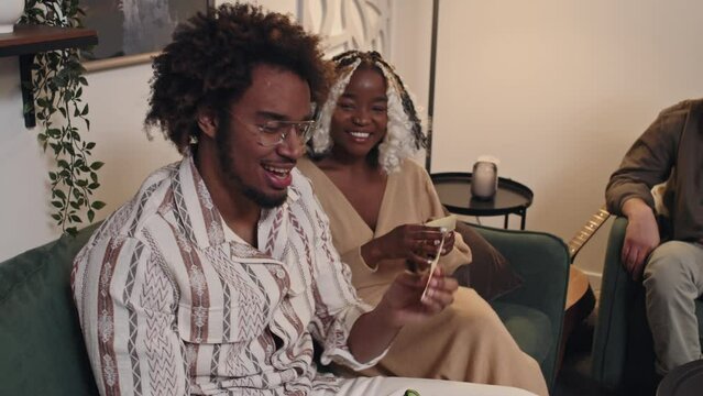 Slow motion of stylish young Black guy sitting in living room with friends posing on camera with guessing game sticker