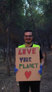 Guardabosque enseñando una pancarta con el mensaje de ama tu planeta, en la montaña, con expresión sonriente. Video vertical 4K.