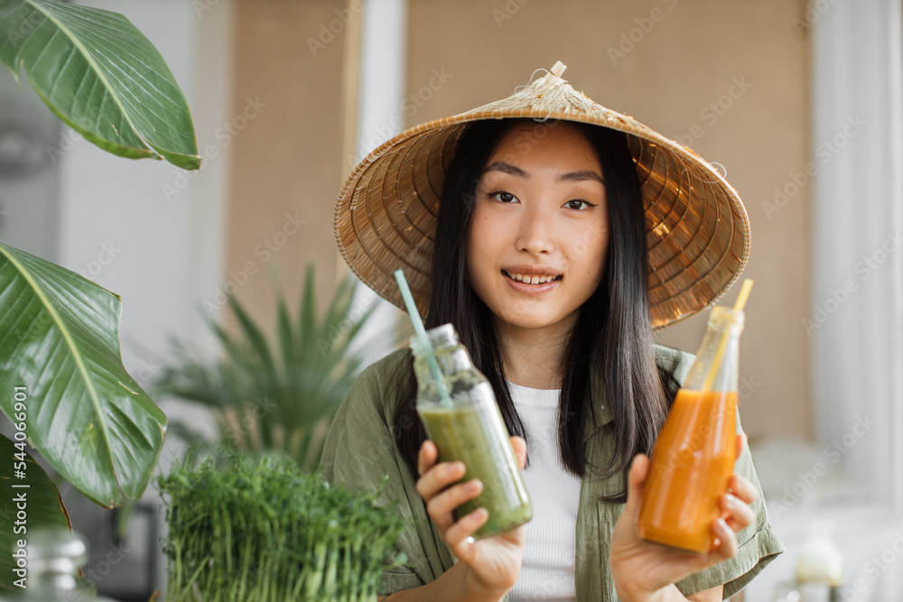 Wall mural Charming young asian vegan woman preparing healthy fresh smoothie for breakfast on tropical exotic kitchen. Attractive female holding in hands two bottle with smoothies. Green healthy food concept.