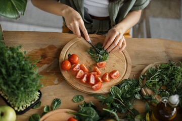 High angle view of hands of young woman chopping arugula leaves, sitting at table in bright exotic studio with many fresh fruits and vegetables are preparing for cooking.