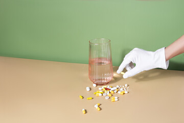 Female gloved hand holding a pill. glass of water and handful of pills are on a table. Well-being...