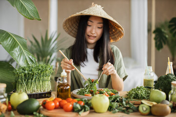 Beautiful asian woman in traditional conical hat stirs fresh healthy vegan salad with fresh vegetables using wooden spoon in tropical exotic kitchen, studio at home, trying a new recipe.